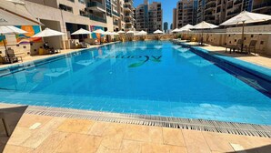 A swimming pool surrounded by multi-story buildings with balconies, outdoor seating, and umbrellas. - Apartment in New Alamein
Furnished Apartment for Rent in Downtown New Alamein (Marsa Matrouh Governorate)