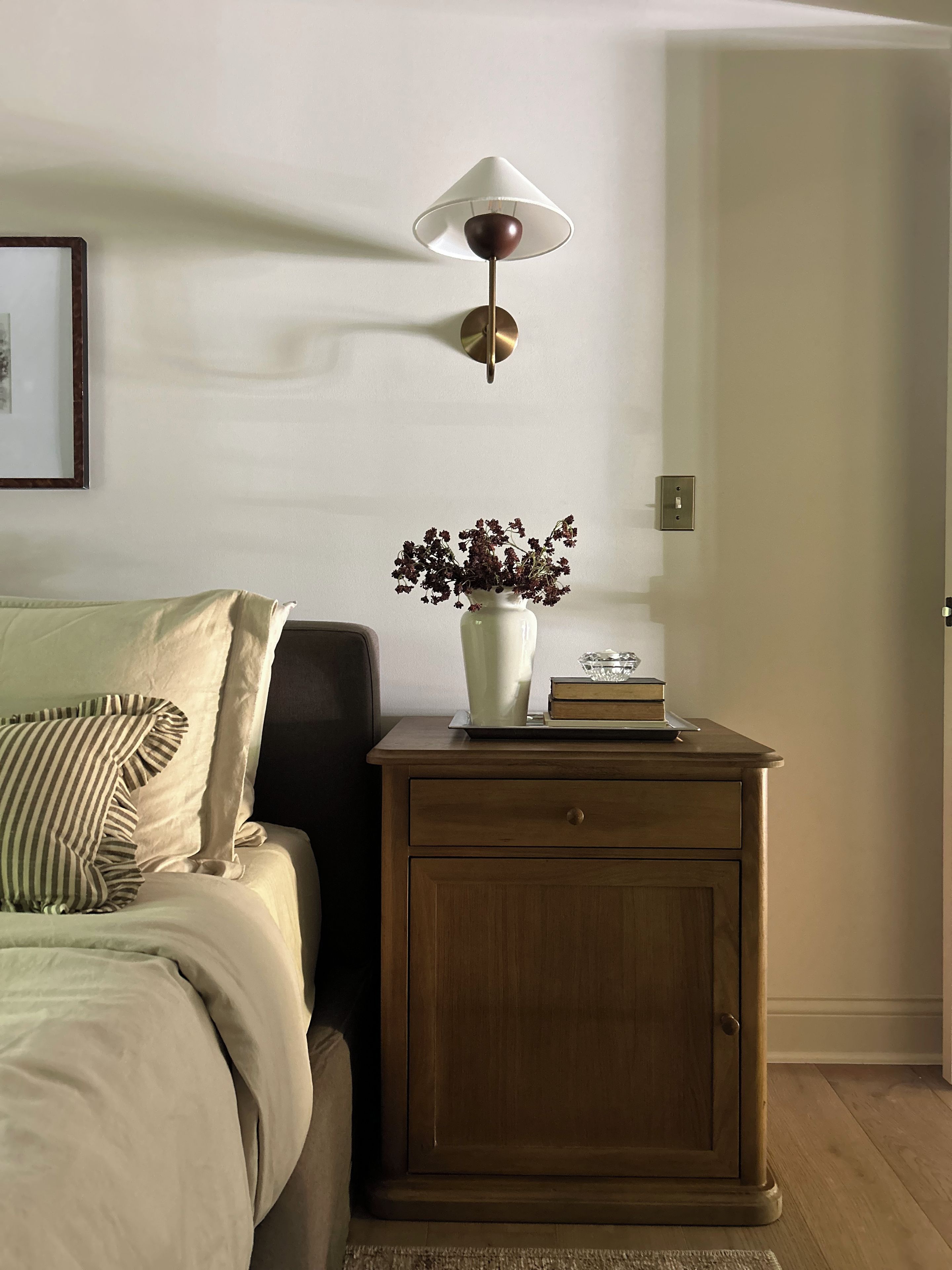 A bedroom with a bed, a bedside table with a vase of flowers, a lamp, and a framed picture on the wall.