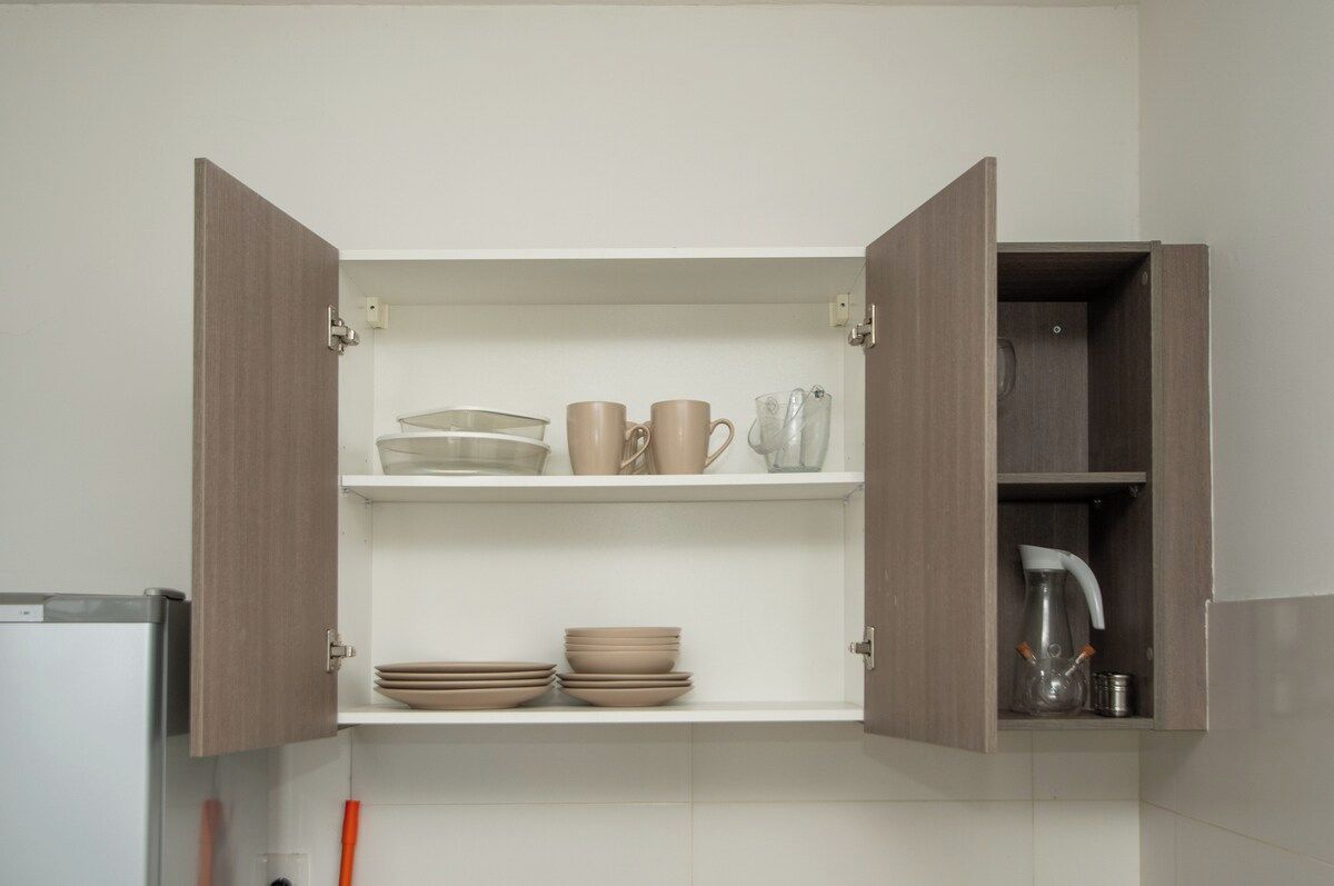 A kitchen with open wooden cabinets containing dishes, cups, and a kettle.