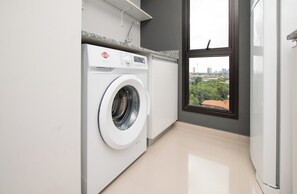 Une petite buanderie équipée d’un lave-linge à chargement frontal, d’une fenêtre donnant sur un paysage urbain et d’un plan de travail avec un évier.