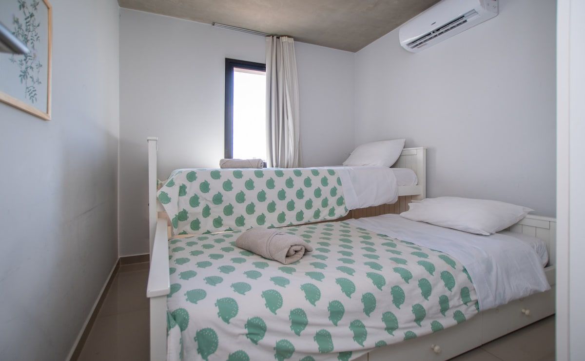Un lit simple avec une couvre-lit à motifs, une fenêtre avec des rideaux et un appareil de climatisation fixé au mur.
