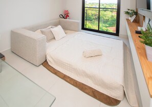 A modern bedroom with a sofa bed, a flat-screen TV, a fire extinguisher, and a view of greenery through a large window. - Zuba 13 1BR | Pool, WiFi & Near Airport (Luque)