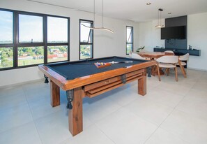 Mesa de bilhar com estrutura de madeira e superfície em feltro preto, área de refeições com cadeiras e mesa, e televisão fixada na parede.