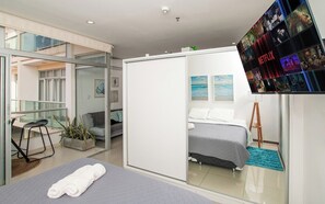 A modern hotel room with a bed, a seating area with a chair and a small table, a large TV screen displaying Netflix content, and a glass partition separating the room from the adjacent space. - Modern Retreat with Pool, Gym and Private Balcony (Asunción)