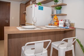 Eine Küchenzeile mit Kühlschrank, Mikrowelle und Spüle. Zwei weiße Sessel mit Armlehnen und ein kleiner Tisch mit einem Weinglas und Tellern.