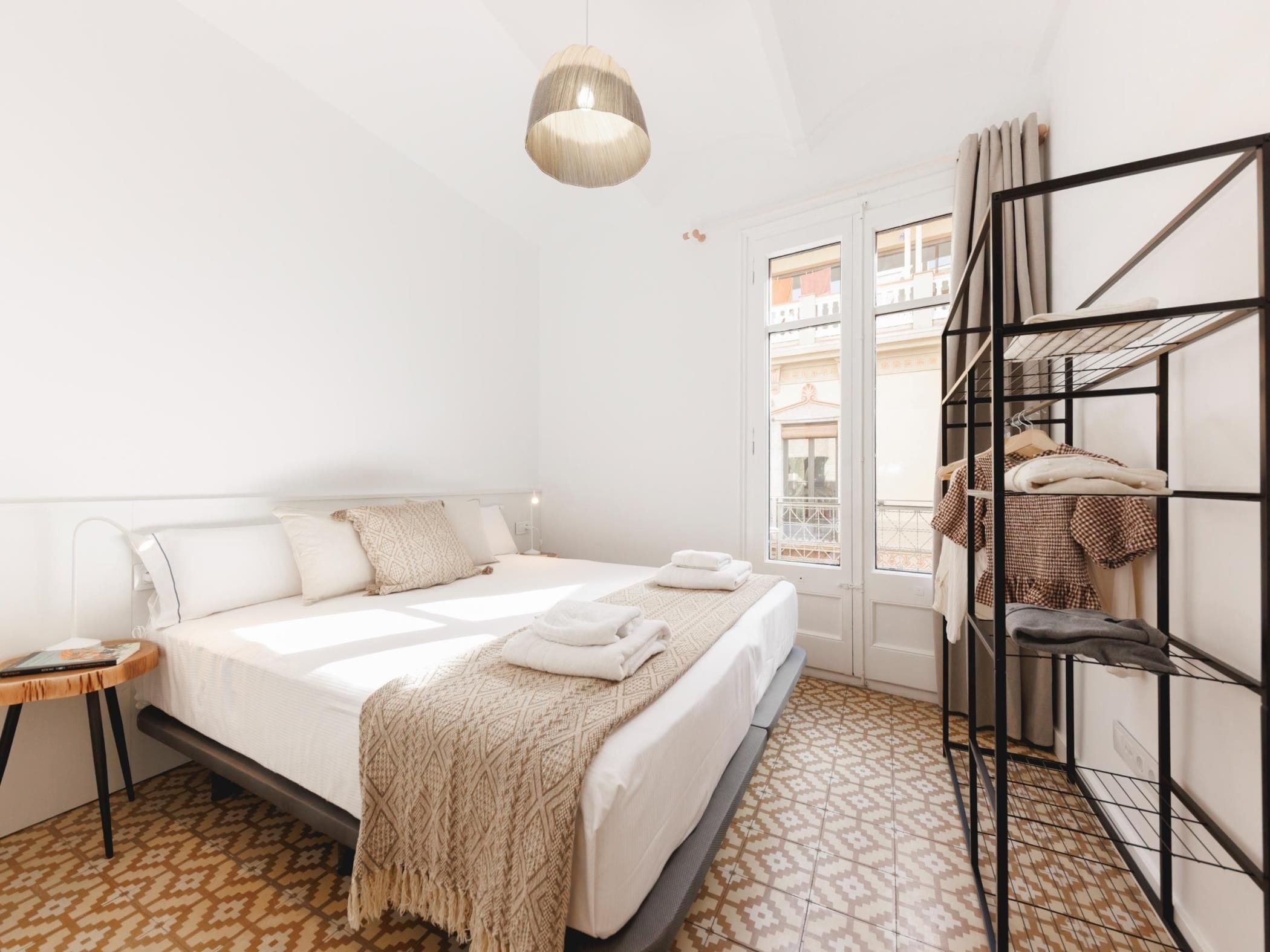 A hotel room with a bed, a bedside table, a hanging light fixture, a window with a view, and a built-in shelf unit.