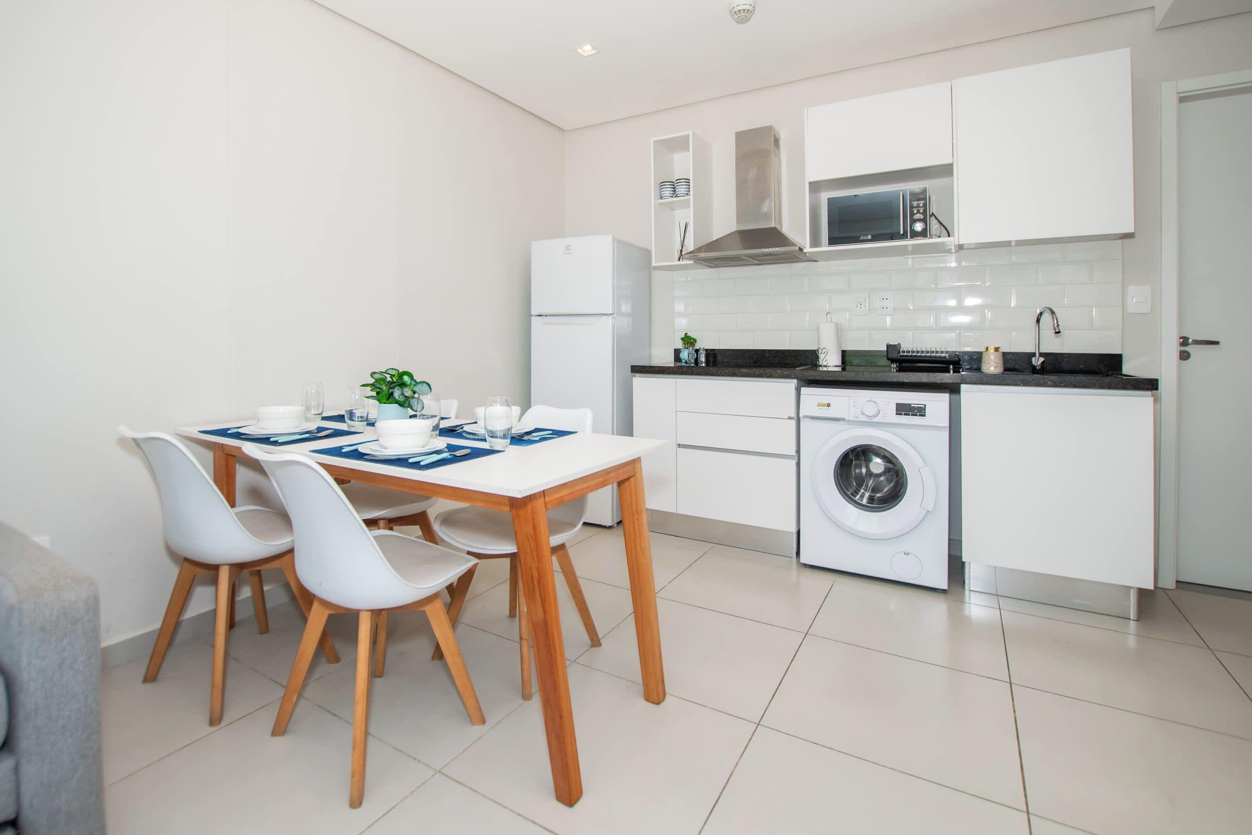 Una cocina moderna con armarios blancos, lavadora integrada y comedor con mesa de madera y sillas blancas.