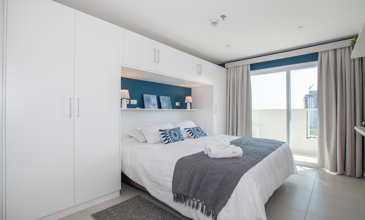 A modern bedroom with a bed, white wardrobe, and a city view. - Work Ready Hub with Gym, Pool&Shopping in Mariscal (Asunción)