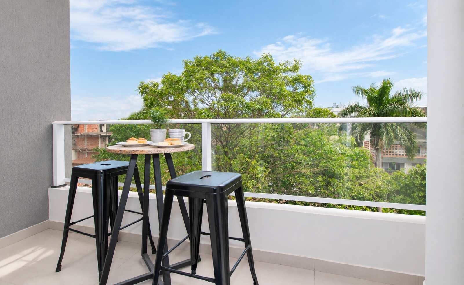 Un balcón con dos taburetes negros, una mesita redonda pequeña y una barandilla blanca, con vistas a zonas verdes y edificios.