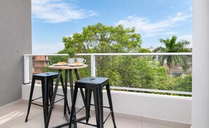 Ein Balkon mit zwei schwarzen Hocker und einem kleinen runden Tisch, einem weißen Geländer sowie Blick auf Grünflächen und Gebäude.