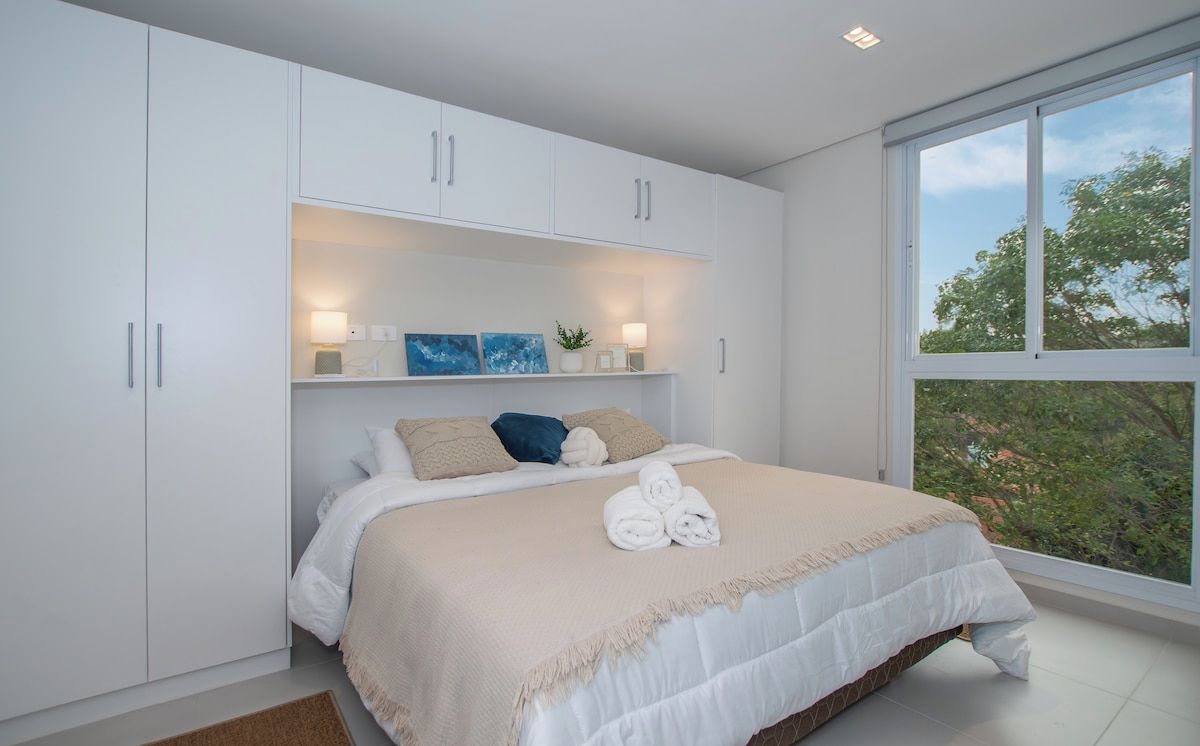 Een slaapkamer met een groot bed, witte kasten, een raam met uitzicht en een plank met decoratieve objecten.