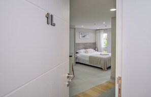 Habitació d'hotel amb un llit, una taula de nit, una finestra amb cortines i una porta amb el símbol de número.
