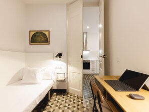 Ein kleines Hotelzimmer mit einem Bett, einem Schreibtisch mit einem Laptop, einem Sessel, einer Küchenzeile und einem gerahmten Bild an der Wand.