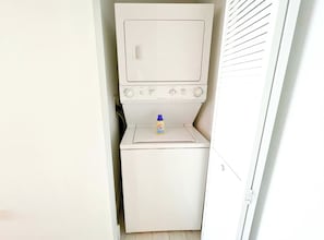 A compact laundry room with a washing machine and dryer, a small shelf with a detergent bottle, and a door with a handle. - 1BR- Intracoastal & Marina - Private Balcony (Hallandale Beach)