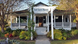 Un edificio con l'indicazione "Gulf Shores Museum", dotato di veranda, scale e un giardino con diverse piante.
