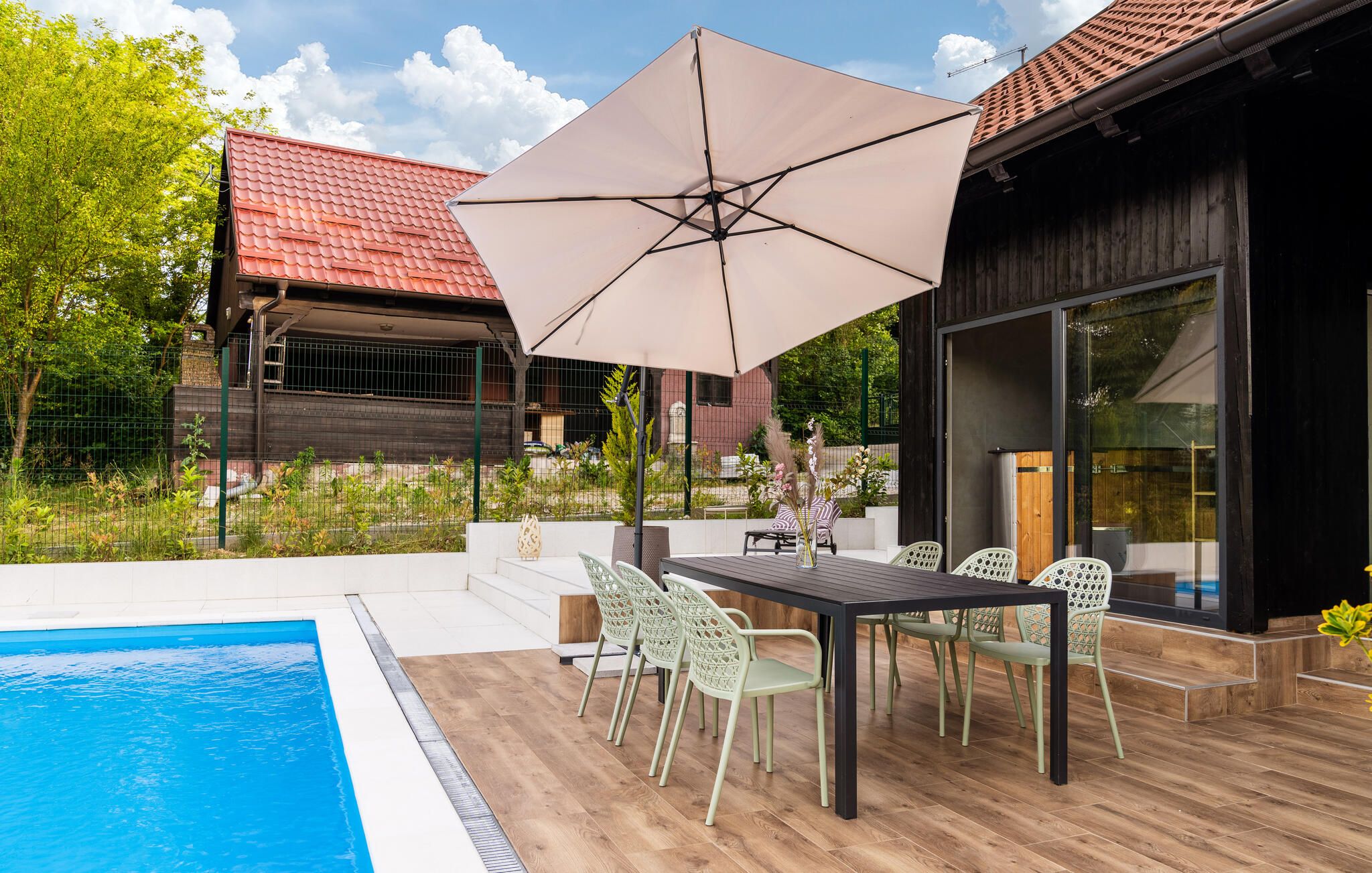 A poolside area with a table and chairs, a large umbrella, and a view of a house with a red roof.