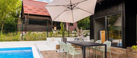 Un espace aménagé au bord de la piscine, avec une table et des chaises, un grand parasol et une vue sur une maison avec un toit rouge.