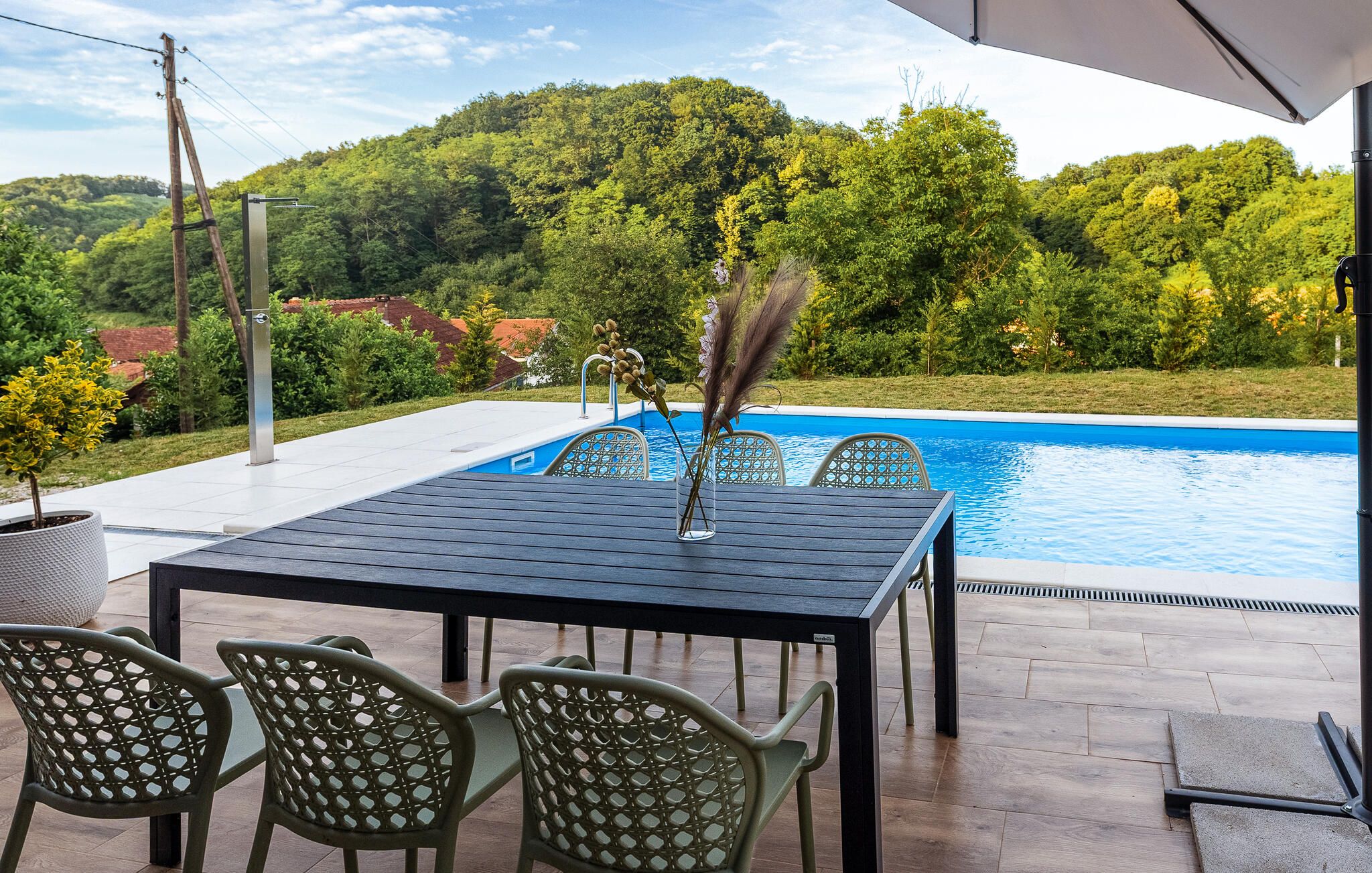 An outdoor pool area with a table and chairs, surrounded by a pool and a view of a forested hill.