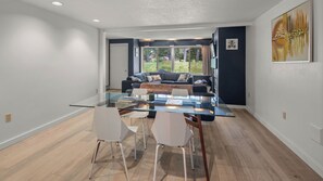 Ein modernes Wohnzimmer mit einem Glastisch, weißen Stühlen, einem dunkelgrauen Sofa und Blick auf einen Garten durch große Fenster.