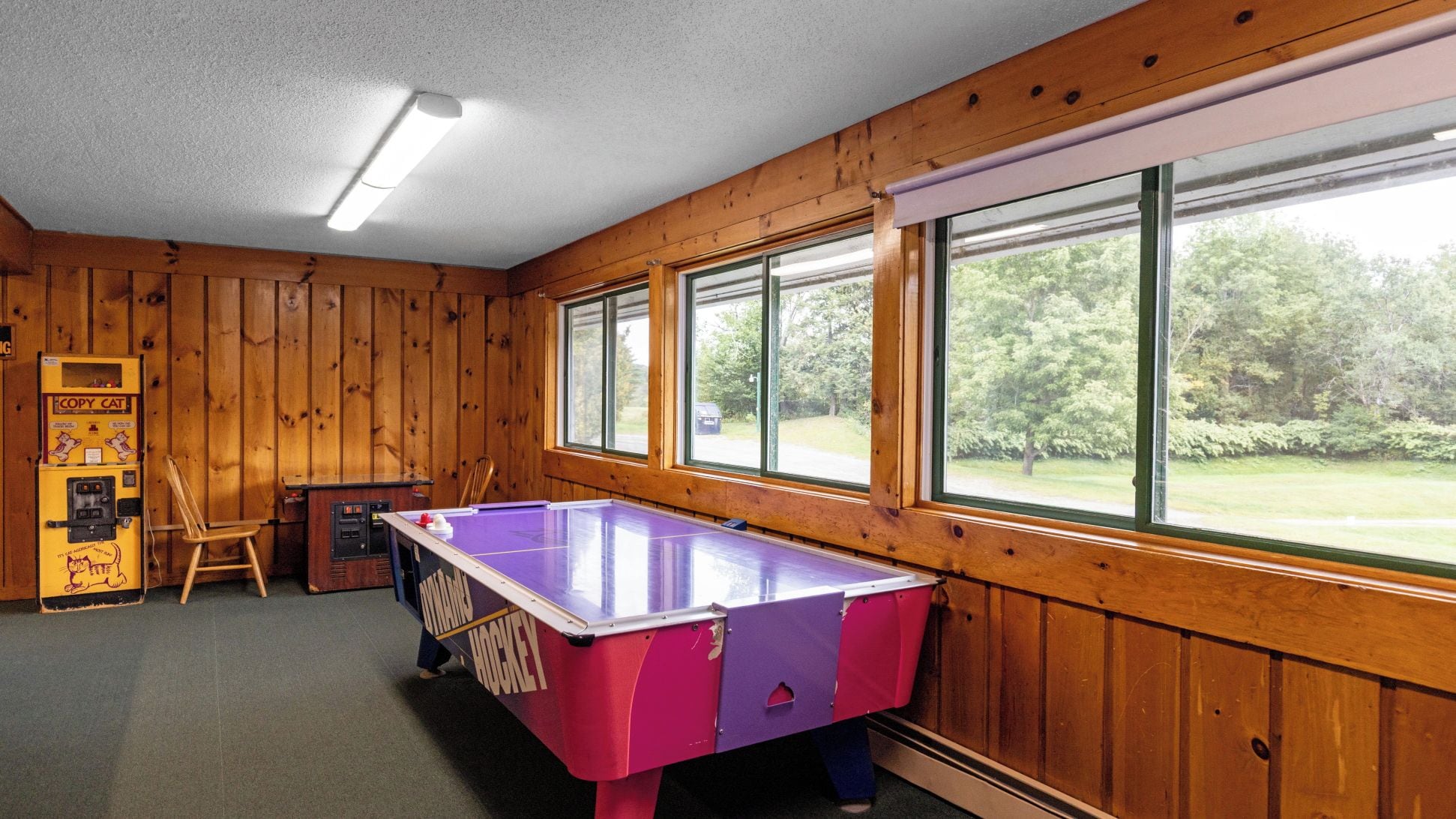 A room with a foosball table, a jukebox, and large windows with a view of greenery.