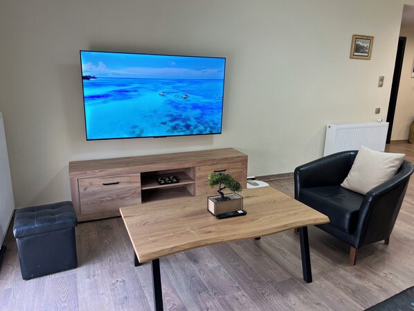 Una sala de estar con un televisor de pantalla plana montado en la pared, una mesa de centro de madera, un sofá de cuero negro y una pequeña planta sobre la mesa.