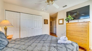 Quarto com cama, penteadeira, ventilador de teto, televisão e porta.