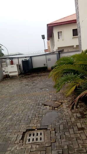 A paved outdoor area with a drainage grate, a white building with a red roof, and a palm plant. - 7 on Dutse Street Hotel (Abuja)