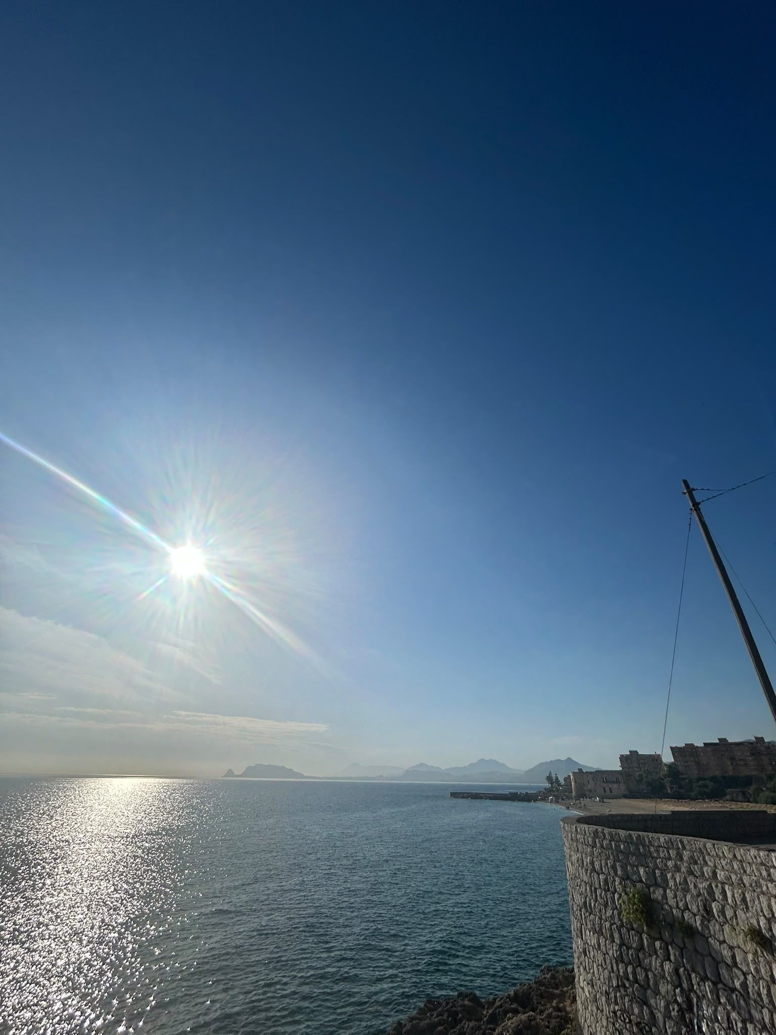 Küstensicht mit blauem Himmel, ruhiger See und einer Steinmauer mit Stacheldrahtzaun.