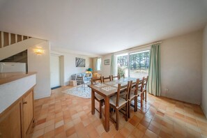A dining area with a wooden table and chairs, a kitchen with a refrigerator, and a staircase leading to an upper level. - Coin de paradis - Grande maison pour 14 pers (Saint-Philibert)