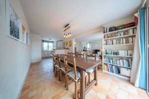 Una àrea de menjador àmplia amb taula i cadires de fusta, una prestatgeria plena de llibres i una cuina al fons.