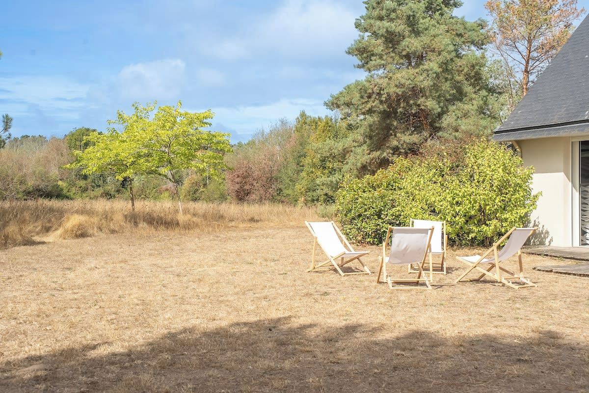 Een groene buitenruimte met drie houten lounge stoelen en een kleine tafel, omgeven door bomen en struiken.