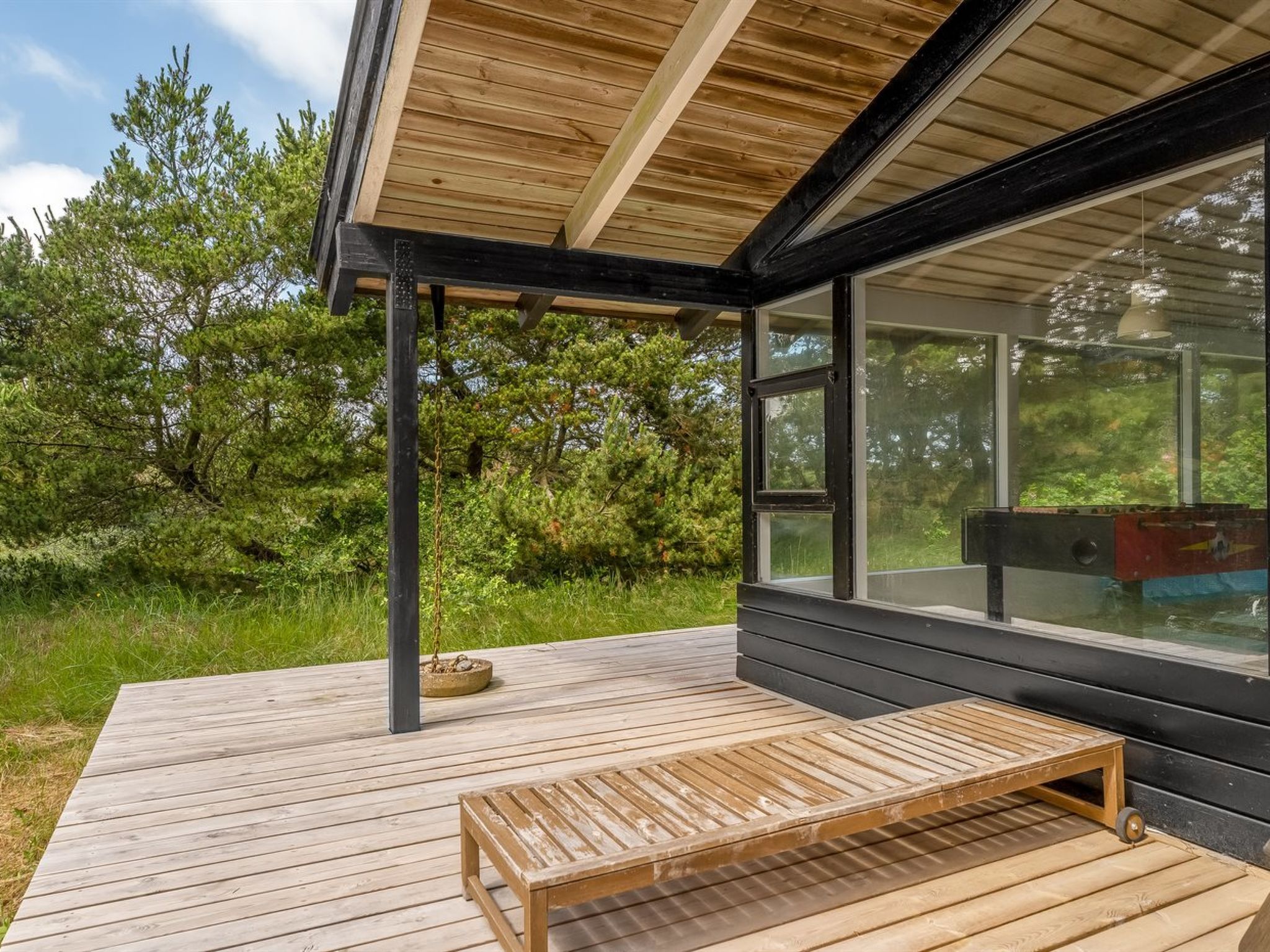 Una terraza de madera con un banco, un cerramiento de vidrio con vistas a la vegetación y un edificio moderno con techo inclinado.