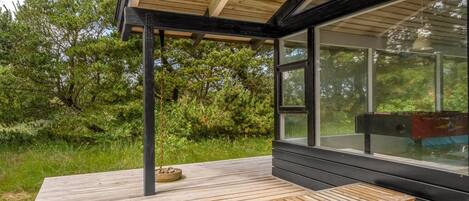 Una terraza de madera con un banco, un cerramiento de vidrio con vistas a la vegetación y un edificio moderno con techo inclinado.
