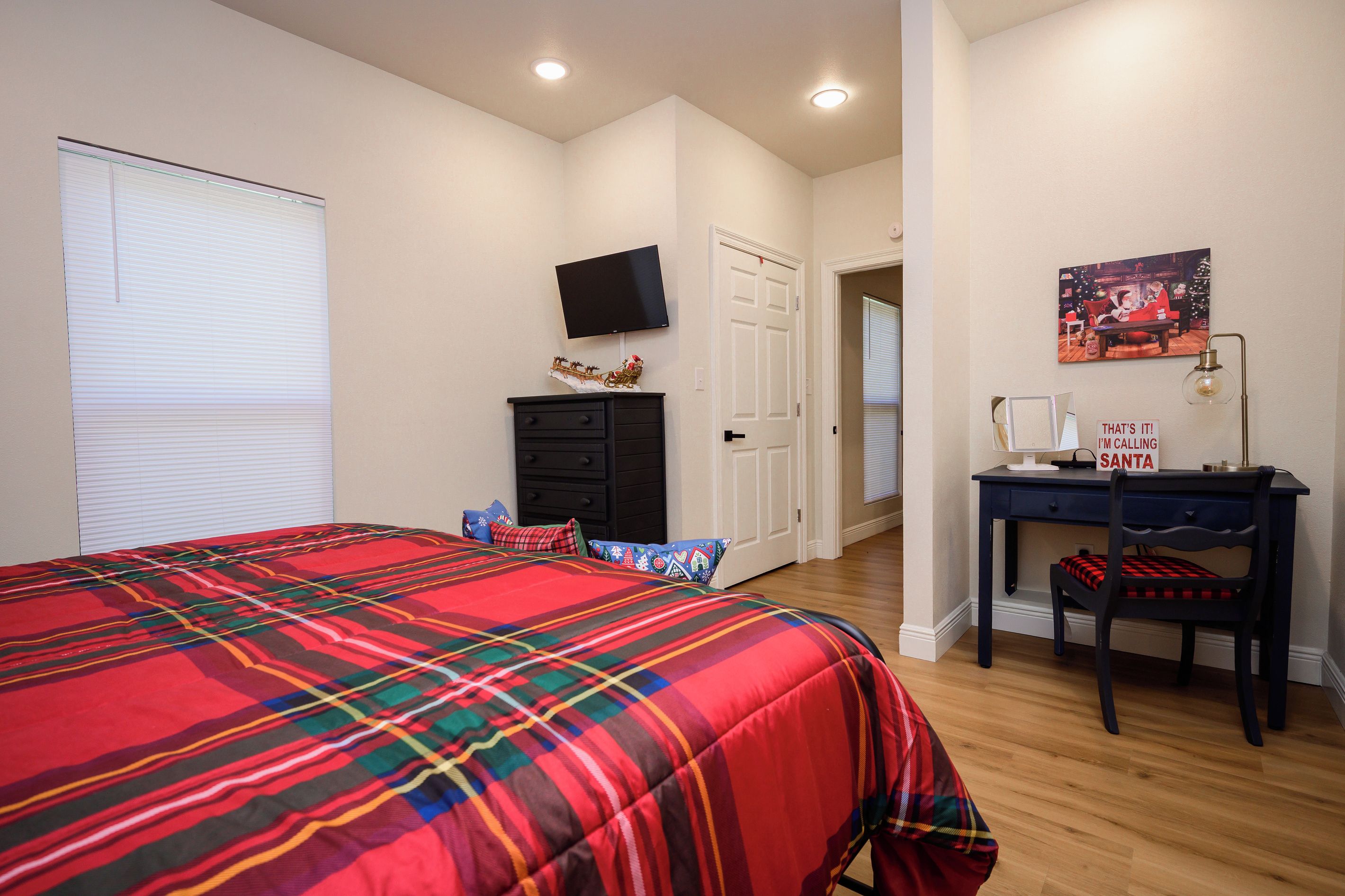 Quarto com cama, cômoda, escrivaninha com cadeira, televisão na parede e porta que dá para outro ambiente.