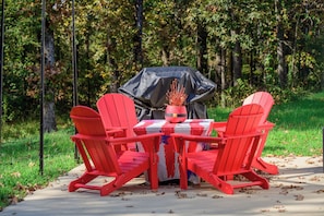 Ein roter Adirondack-Stuhl steht um einen Tisch mit Grill und Sonnenschirm in einer bewaldeten Außenanlage.