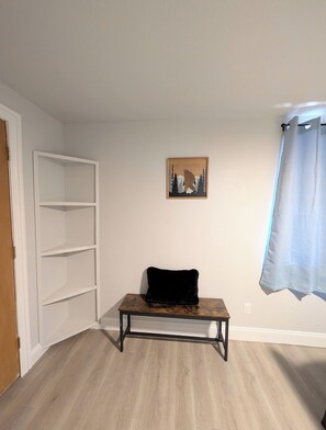Una habitación con piso de madera, un cojín negro sobre una mesita, una puerta blanca y un cuadro enmarcado en la pared.