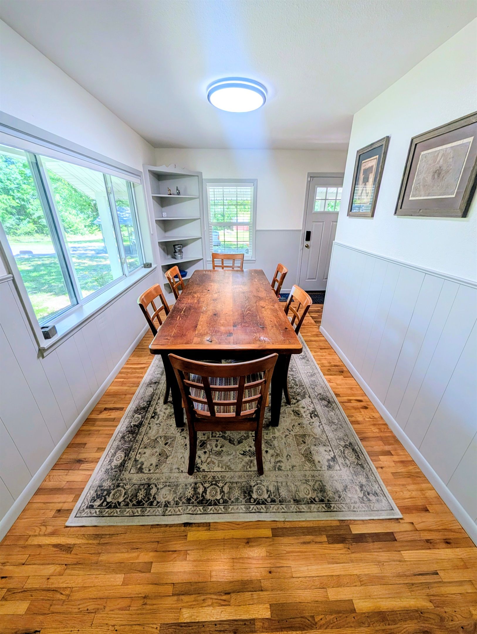 Une salle à manger avec une table et des chaises en bois, un tapis à motifs et une vue sur la verdure à l’extérieur.