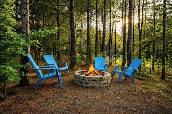 A forest setting with a stone fire pit and three blue Adirondack chairs arranged around it. - Pretty in the Pines- *Near WPG* (MENA)