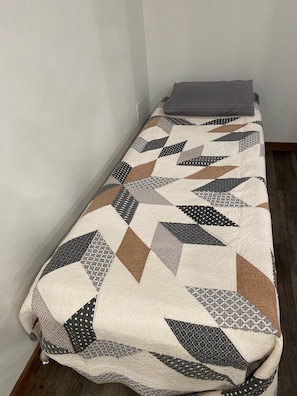 A single bed with a patterned bedspread in a room with plain walls and wooden flooring. - Whispering Pines Guest Cabin near Wolf Pen Trails! (Mena)