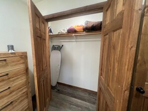 A narrow walk-in closet with wooden doors, a hanging iron, and a shelf with folded clothes. - Whispering Pines Guest Cabin near Wolf Pen Trails! (Mena)