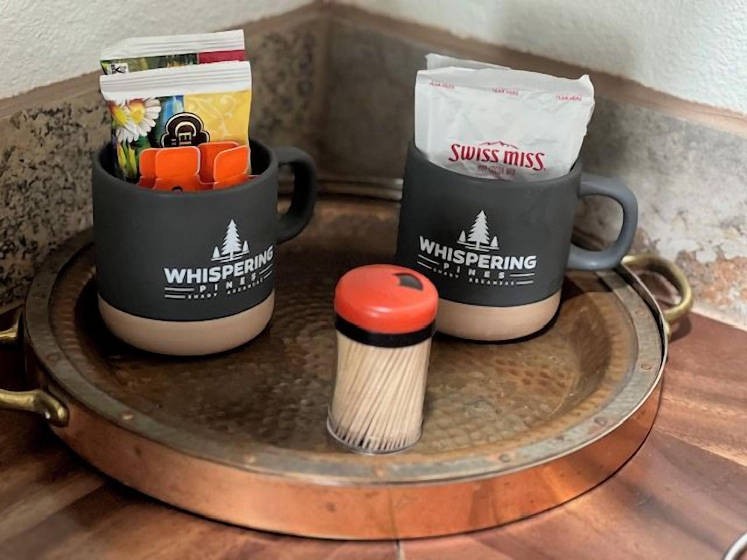 Two mugs with the 'Whispering Pines' logo, a red container with a white lid, and a tray with a copper base.