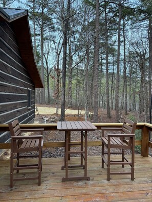Una terraza de madera con dos mesas de picnic y bancos, rodeada por un bosque.