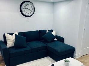 A living room with a teal sectional sofa, a white rug, a white coffee table with a plant, and a wall clock. - Private Bsmt Apt W/Separate Entry- 1 Min to Subway (Oak Park)