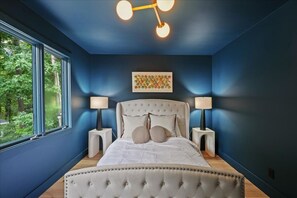 A bedroom with a large bed, two bedside tables, a ceiling light fixture, and a window with a view of trees. - The SB Gallery House Unapologetically Different (Raleigh)