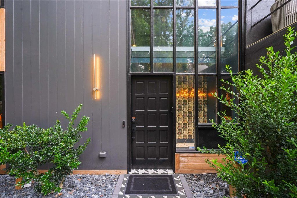 A modern building with a dark facade, a black door, and a small garden area with plants and a doormat.