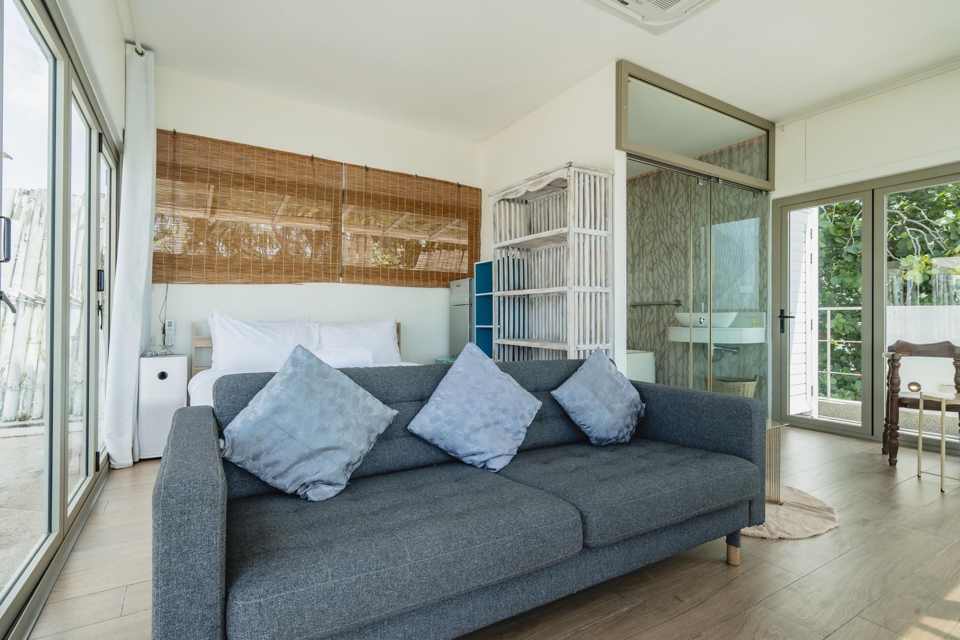 A modern bedroom with a grey sofa, a bedside table, a large window, and a sliding glass door leading to a balcony.