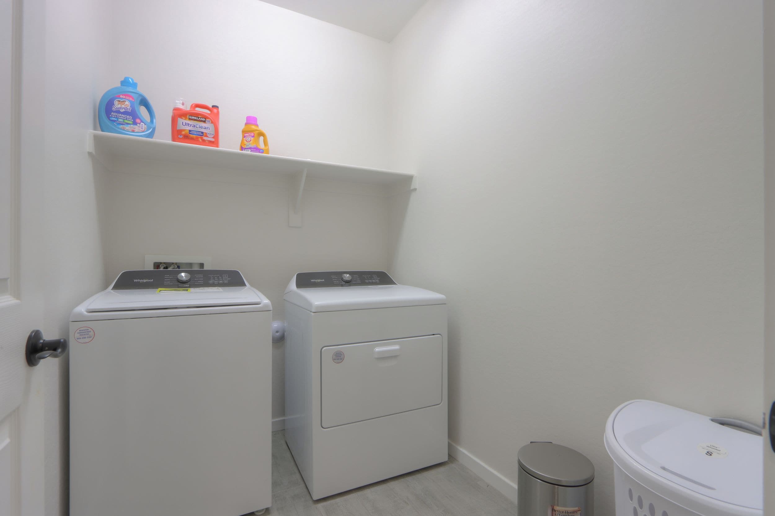 A laundry room with two front-loading washers and a shelf above containing cleaning supplies.