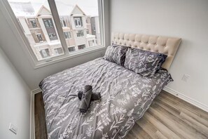 A bedroom with a tufted headboard, a grey floral bedspread, and a view of buildings through a window. - Luxury 3BR Retreat in South Barrie • Parking (Barrie)