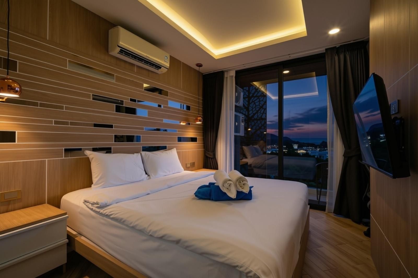 A modern hotel room with a large bed, a flat-screen TV, and a view of the city at dusk.