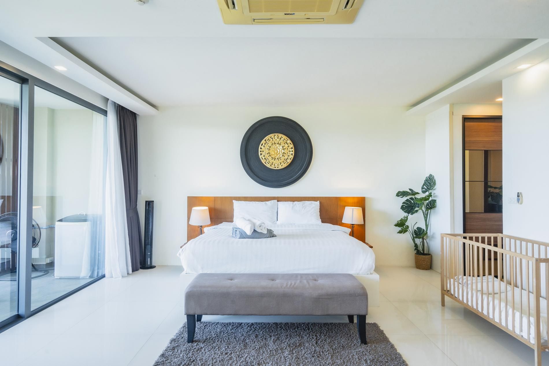Une chambre d’hôtel avec un lit, des lampes de chevet, un objet décoratif rond accroché au mur, un banc gris, une plante en pot et un berceau.